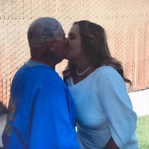 A man with extensive tattoos on his head and neck, wearing a blue shirt, kisses a woman in a white top and pearl necklace against a brick wall.