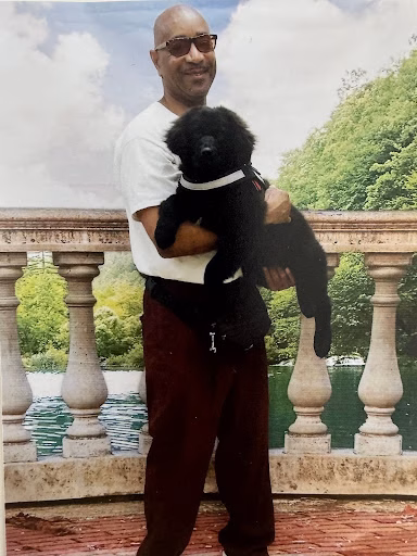 Jeffery, a bald man with sunglasses, holds a small black dog in a white shirt and maroon pants on a stone balcony overlooking a lake.
