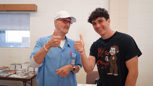 Two men, one older and one younger, both wearing white wristbands, give thumbs up in a room.