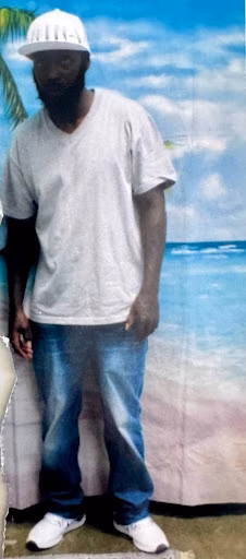 Randy, a man with a beard, wearing a white cap, grey t-shirt, and blue jeans, stands in front of a beach backdrop.