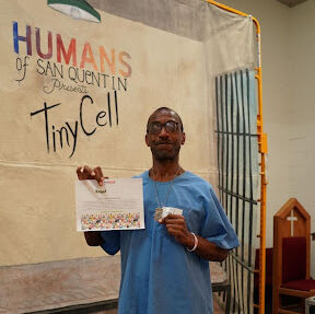 A man in blue scrubs holds a certificate in front of a banner reading 'Humans of San Quentin Presents Tiny Cell'.