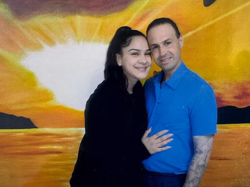 A man and a woman posing together in front of a vibrant, sunburst-themed mural.