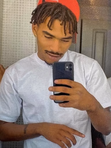 Treavion, a man with dreadlocks and a red cap, takes a mirror selfie with a smartphone.