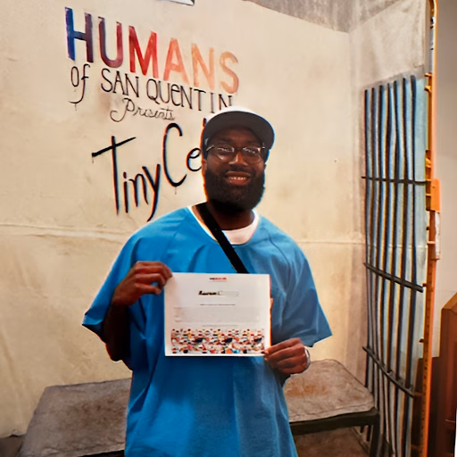 Aaron, wearing a blue shirt and cap, smiles while holding a certificate in front of a wall that reads "Humans of San Quentin Presents Tiny Celebrities".