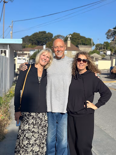 Three people, Bruce and two women, standing together on a sidewalk in a residential neighborhood.