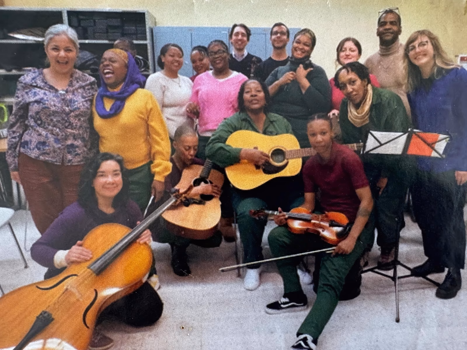 A diverse group of musicians, including a woman playing a cello, a man with an acoustic guitar, and a young person with a violin, posing together in a room.