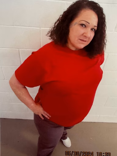 Jimidenise "Niesey" wearing a red shirt and brown pants, posing against a white brick wall.