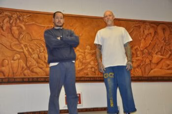 Two men stand in front of a large, detailed relief mural depicting a battle between angels and dragons. The mural includes a decorative border with religious text.