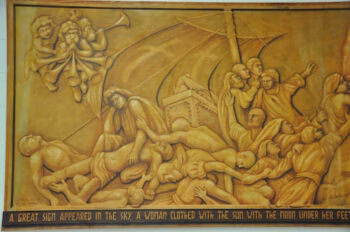 A golden relief mural depicting a dramatic biblical scene with figures in distress, a ship, and angels above, with a descriptive text at the bottom.