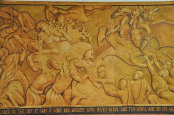 {"alt_text": "A detailed bas-relief mural depicting a chaotic battle scene with numerous human figures and a large, multi-headed dragon, set against a textured, earthy-toned background.", "title": "Mural piece 3", "caption": "A section of a large-scale bas-relief mural showing a dramatic battle scene.", "description": "This image displays a detailed bas-relief mural, identified as piece 3, featuring a dynamic and chaotic battle scene. The artwork is carved into a textured, earth-toned surface and depicts numerous human figures in various states of action, including combat and flight, engaged in a struggle with a large, multi-headed dragon. A partial inscription at the bottom of the mural reads, 'SCARED IN THE SKY IT WAS A HUGE RED DRAGON WITH SEVEN HEADS AND TEN HORNS AND ON ITS WE...'}