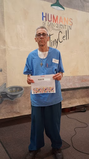 Robert R stands in front of a 'Humans of San Quentin' mural, holding a certificate.