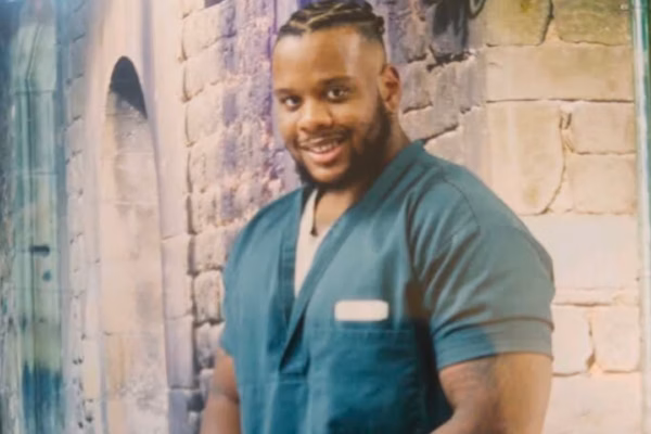 Portrait of Elijah, 31, a man with braided hair and a beard, wearing teal scrubs and smiling, standing in front of a stone wall.