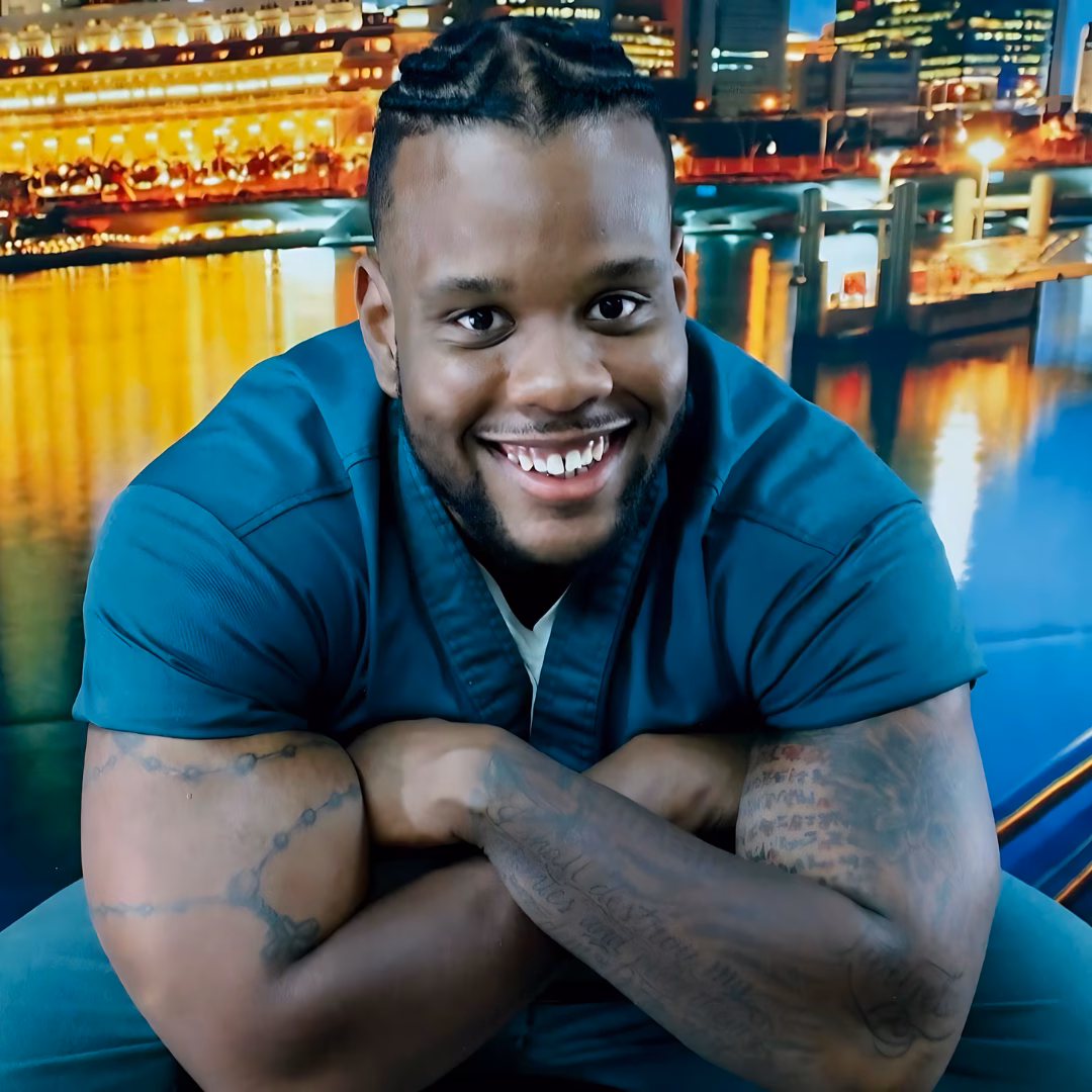 Portrait of Elijah, 31, smiling with arms crossed, wearing a teal shirt against a vibrant city waterfront night background.