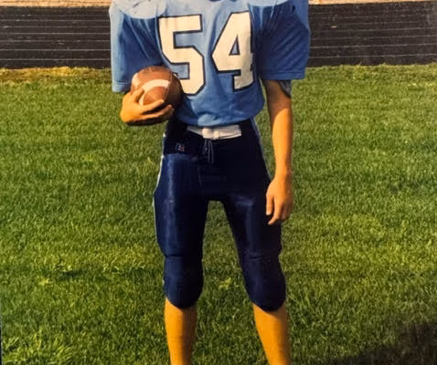 unnamed-4 Kyle, 40, a football player in a blue jersey with the number 54, stands on a green field holding a football with a helmet on the ground.