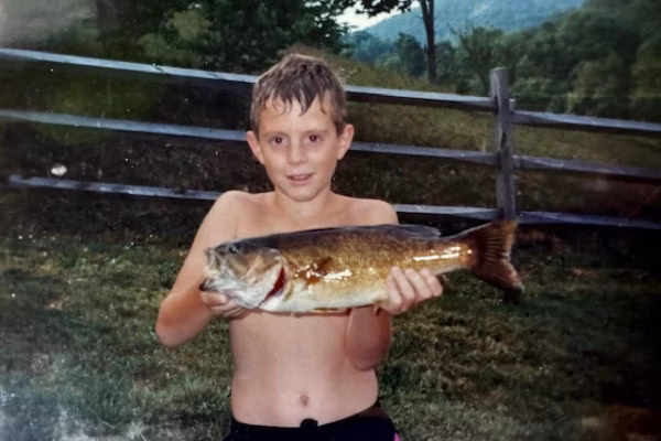 unnamed-5 A young boy holds a large fish in front of a wooden fence with a mountainous landscape in the background.