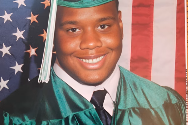 A portrait of Elijah, 31, wearing a green graduation cap and gown, smiling in front of an American flag.