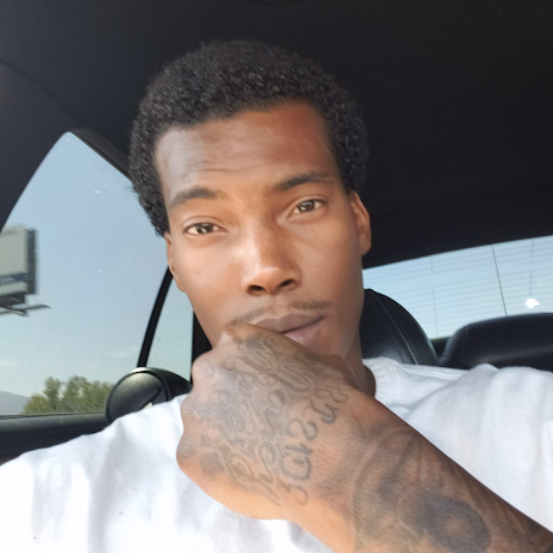 Greg Maurice inside a car, wearing a white shirt and displaying a tattooed hand near his mouth.