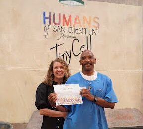 A man in blue scrubs and a woman in black hold a certificate in front of a wall reading 'Humans of San Quentin Presents Tiny Cell'.