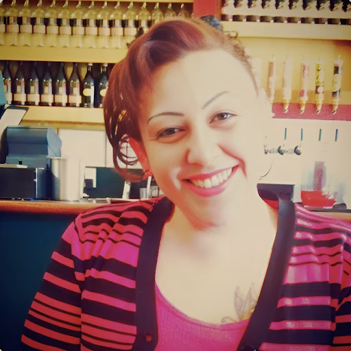 Bobbie smiles warmly in a pink and black striped cardigan, with a tattoo on her chest, in a bar setting.