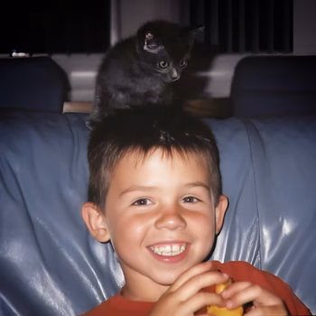 A young boy with a black cat perched on his head, both looking towards the camera.