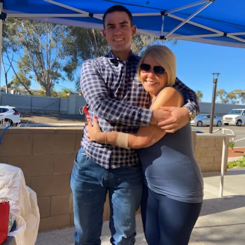 A man and a woman, Casey, 29, share a warm embrace under a blue canopy on a sunny day.