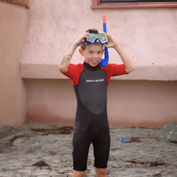 Casey, 29, a young boy in a black and red wetsuit with 'GOGO GLOVE' text, adjusts a snorkel mask on a sandy beach.