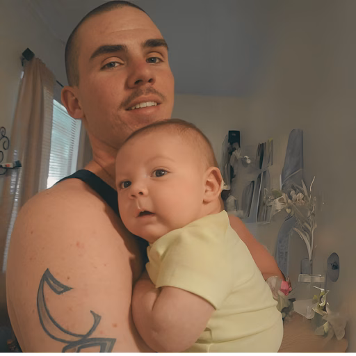 A man named Casey with a tattoo on his arm holds a baby in a yellow shirt.