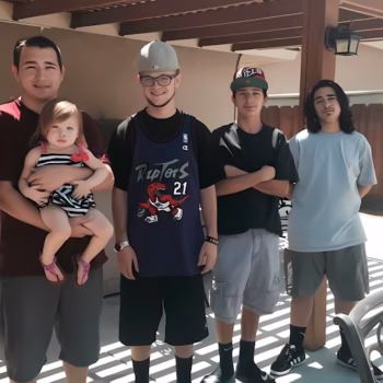 Four men and a baby standing together under a wooden pergola. The man on the far left holds a baby in a striped dress. The second man wears a blue Raptors 21 jersey and a grey cap. The third man wears a black shirt and a red and black cap. The fourth man wears a light blue t-shirt.