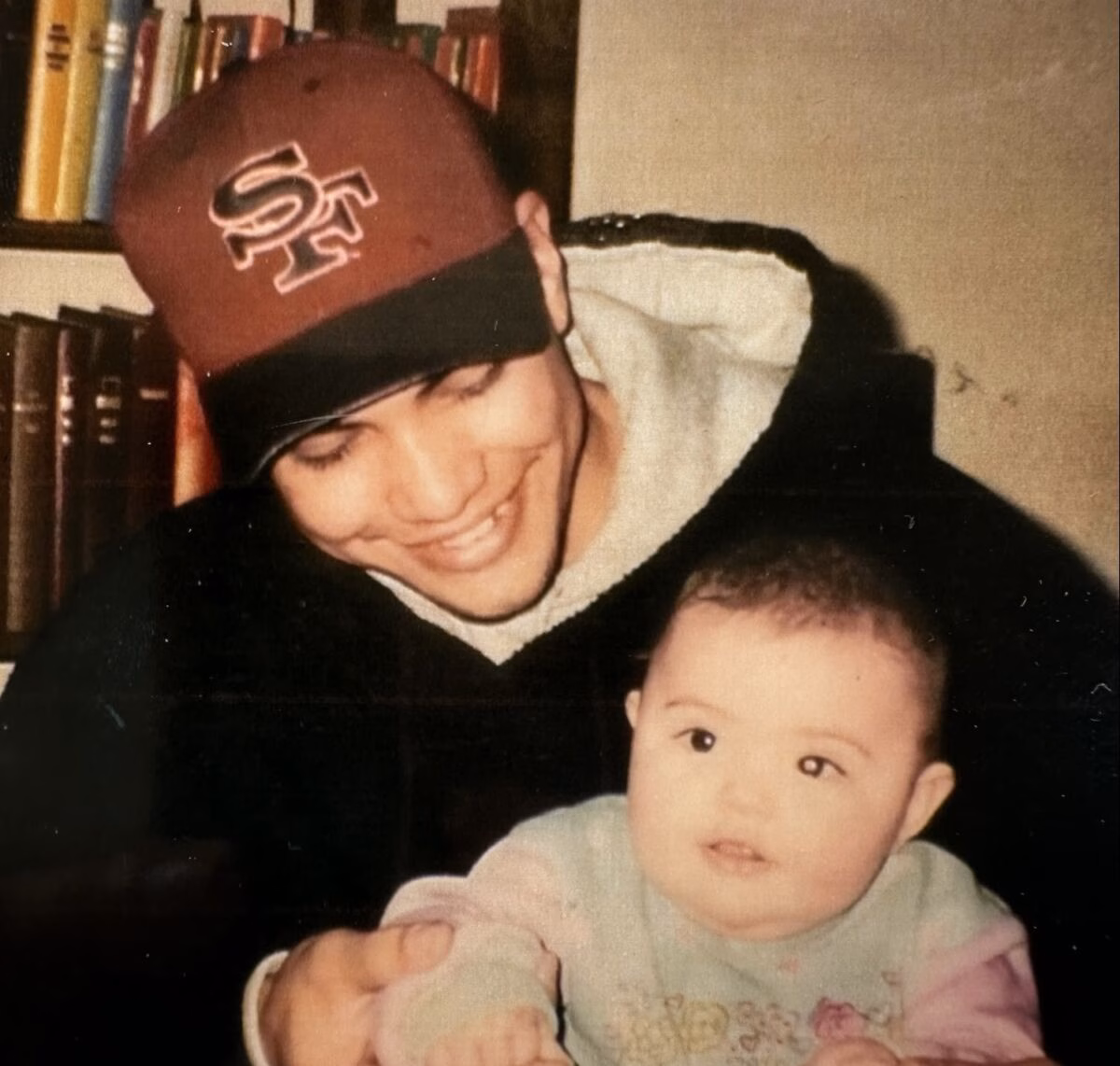 A man wearing an SF baseball cap and a black hoodie smiles while holding a baby in a light-colored outfit.