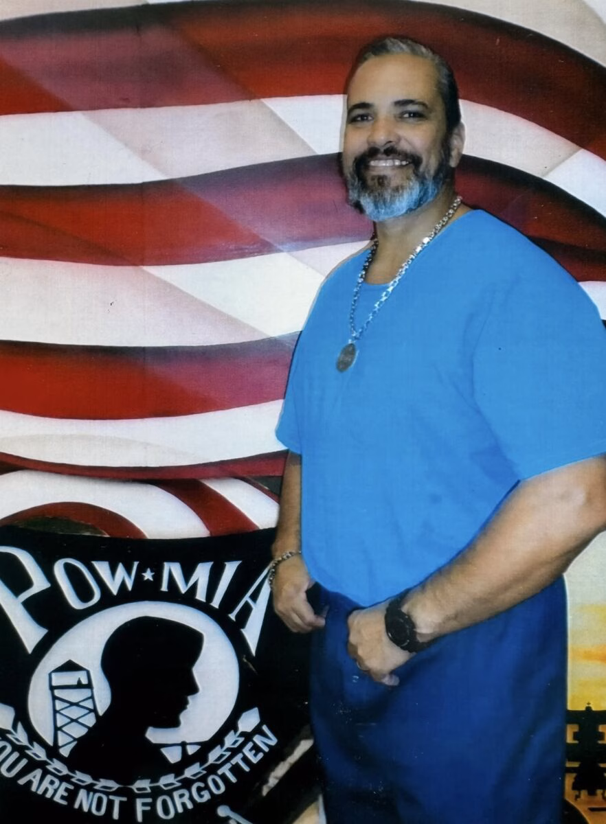 A man with a beard in a blue shirt stands in front of a POW-MIA banner.