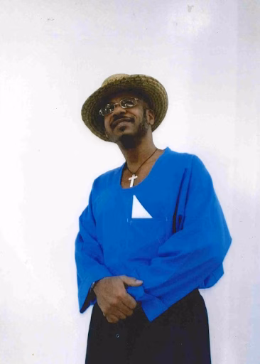 Marvalyon, wearing a straw hat and glasses, smiles while wearing a blue robe with a white triangle and a cross necklace.