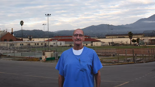 Michael stands in a blue scrub top in front of a sports complex with mountains in the background.