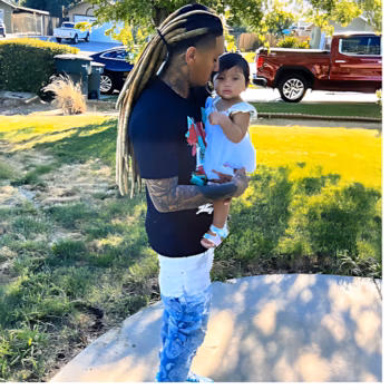 Ray, 32, holding his young daughter in a white dress on a sunny day in a suburban neighborhood.
