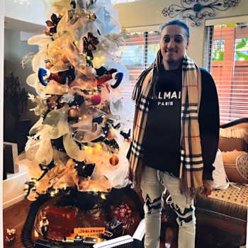 A person with long hair and a Balmain Paris shirt stands next to a white and gold Christmas tree decorated with lights and ornaments.