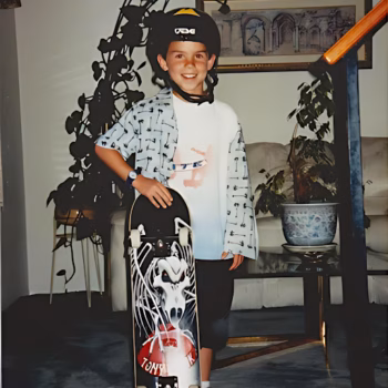 Casey, 29, smiling while holding a skateboard with a skull graphic, wearing a helmet and patterned shirt.