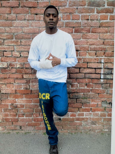 A man named Darius stands against a red brick wall, wearing a white long-sleeved shirt and blue sweatpants with yellow text.