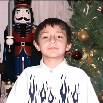 A young boy with a white shirt featuring blue flames stands in front of a large nutcracker soldier and a decorated Christmas tree.
