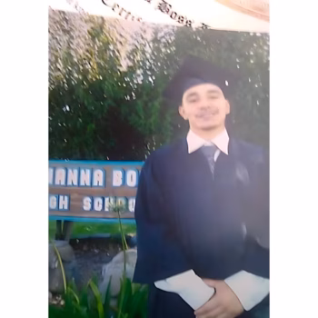 A young man in a graduation cap and gown stands in front of a sign for Gianna Bough High School.