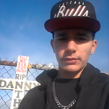 A young man wearing a Chicago Bulls cap and black hoodie stands in front of a chain-link fence with a sign that reads 'Till we see you again RIP DANNY'.