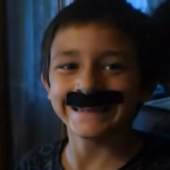 A young boy with a black mustache sticker on his face, smiling.