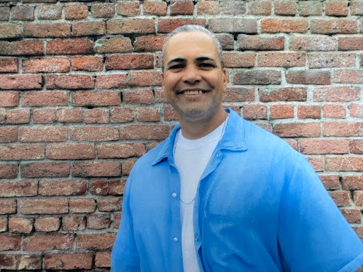 Robert, a man with short grey hair and a beard, smiles at the camera while wearing a blue shirt over a white t-shirt, standing in front of a red brick wall.