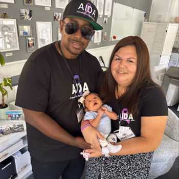 A man and a woman, both wearing black AIDA t-shirts, hold a baby in a well-lit room.