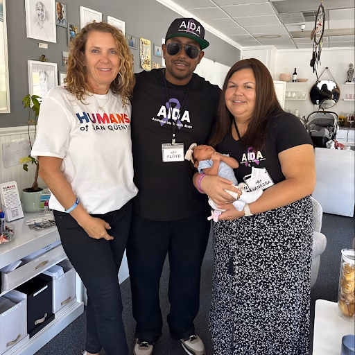 Three people, including a man named Floyd, stand together in an office. Floyd wears a black AIDA t-shirt and a matching cap, holding a baby. The woman on the left wears a white 'Humans of Ian Quint' shirt.