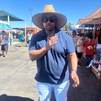 Floyd10, a man in a wide-brimmed hat and sunglasses, gestures while standing in a sunny outdoor market.