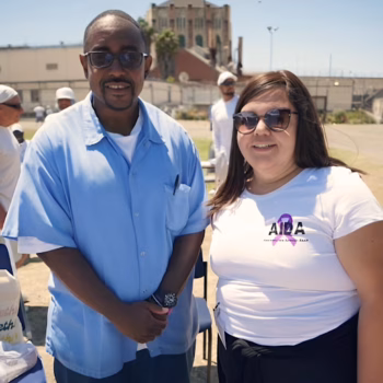 A man in a light blue shirt and a woman in a white t-shirt with the AIDA logo and a purple ribbon stand together outdoors.