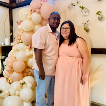 A man and a pregnant woman in matching peach outfits stand together in front of a large balloon arch.
