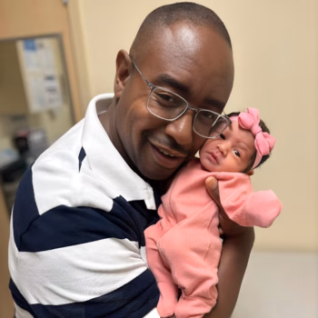 A smiling man in a striped polo shirt holds a newborn baby girl wearing a pink outfit and bow.