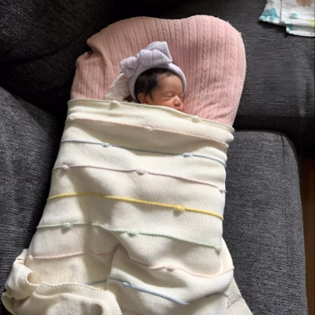 A sleeping newborn baby wrapped in a pastel-colored knitted blanket and wearing a striped bow headband, resting on a pink pillow on a dark grey couch.