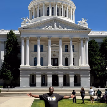 A man in a black T-shirt stands with arms outstretched in front of the white, classical-style California State Capitol building under a clear blue sky.