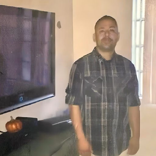 A man named Irwing stands indoors in a plaid shirt next to a television.
